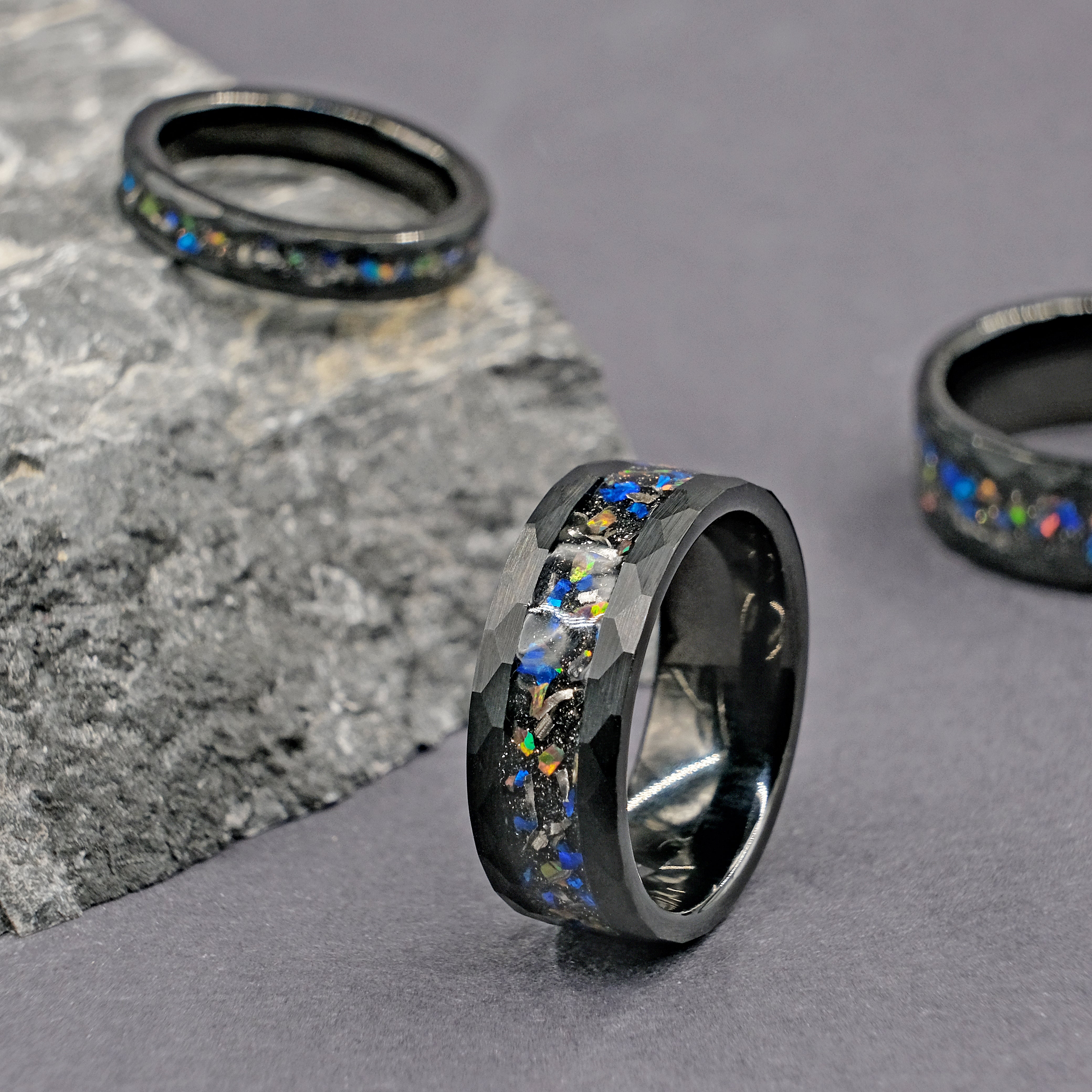 Set of Meteorite Black Tungsten Opal Rings placed on top of a rock, displaying the rugged beauty of the hammered tungsten and the vibrant opal inlay.