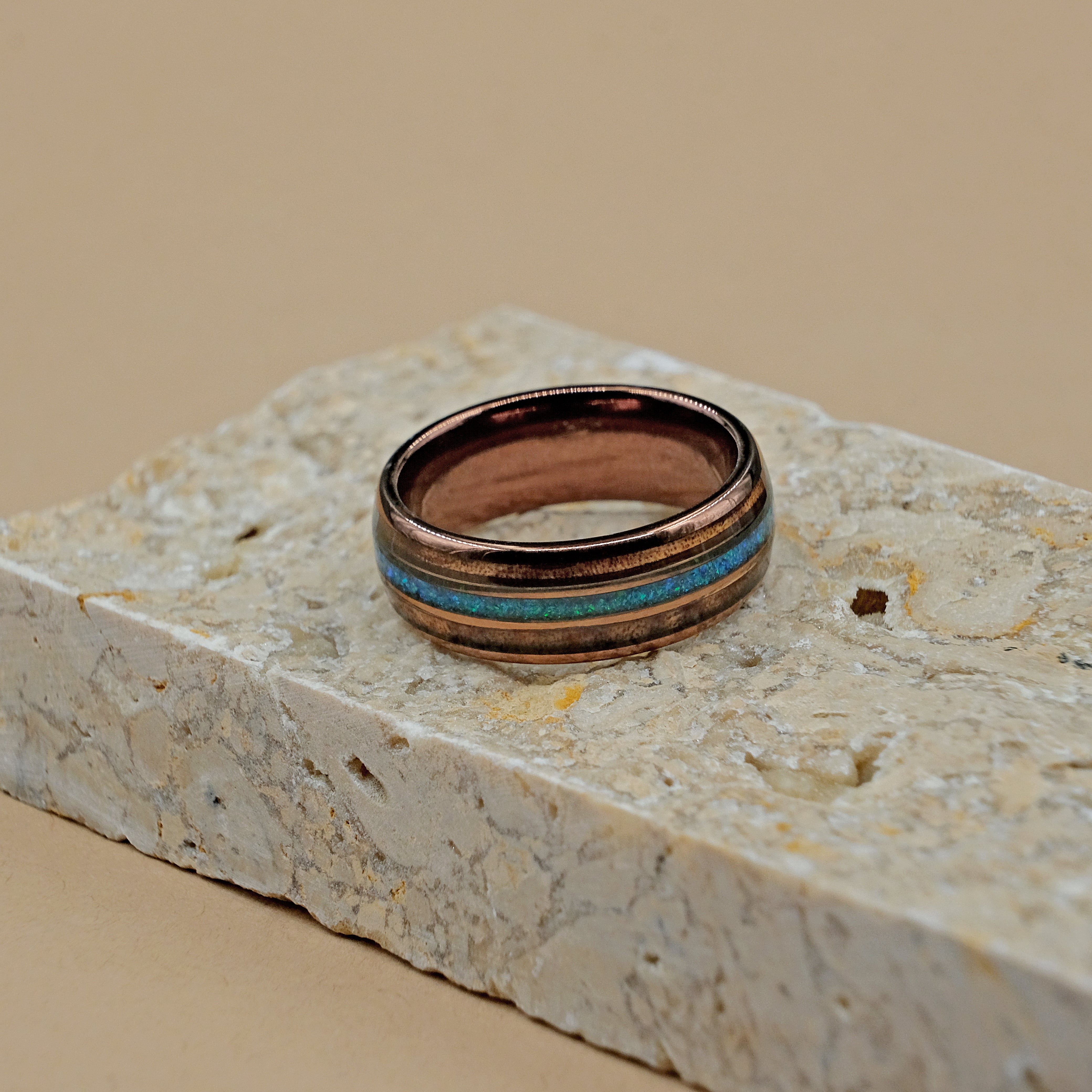 Hawaiian Koa Wood Wedding Ring placed next to a rock, showcasing the intricate inlay of Hawaiian Koa wood and Bello Opal, set in an 18k rose gold-dipped tungsten carbide band.